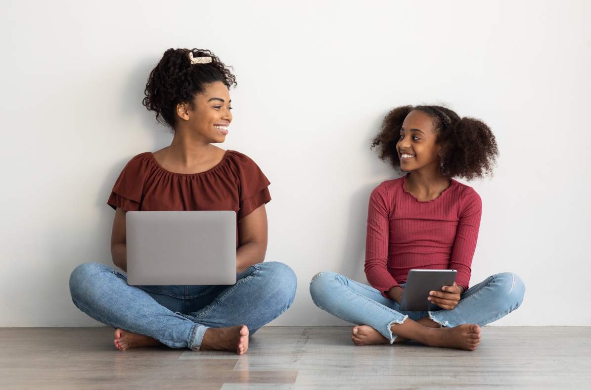 mother sitting with her laptop smiling at daughter beside her on her tablet